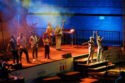 Dieses Bild ist seitlich von der Bühne aufgenommen. Die Band scheint sich zu verabschieden, Die zwei Sängerinnen posieren auf der Treppe, die restlichen 6 Musiker stehen mit ihren Instrumenten auf der Bühne.