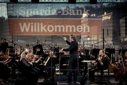 Das Bild zeigt ein Orchester mit ihrem Dirigenten vorne dran. Sie stehen auf der Bühne des Hafen Sommers.