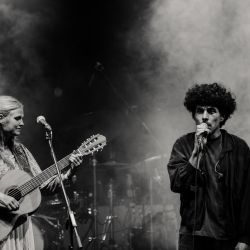 Das schwarz-weiß Bild zeigt links eine Frau mit Gitarre. Rechts einen Mann mit Mikrofon, welcher gerade singt. Die Atmospähre ist durch Nebel sehr mystisch.