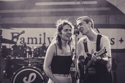Das Bild zeigt zwei Musiker, die sich ein Mikrofon teilen. Links eine Frau und rechts ein Mann mit Gitarre.