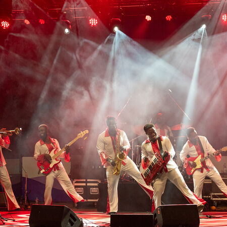 Das Bild zeigt eine Band von 6 personen alle spielen ein Instrument und sie stehen auf einer Bühne. 4 Männer sind in der gleichen Pose, un welcher sie auf dem rechten Bein stehen und das Linke in die Luft heben.