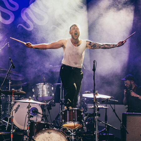 Der Schlagzeuger steht auf seinem Stuhl, er hat beide Arme mit seinen Drumsticks zur Seite ausgestreckt.