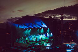 Die Hafensommer Bühne in der Dämmerung. Sie ist blau und grün beleuchet. Das Bild ist sehr dunkel. Auf der Bühne steht eine Band mit 6 Musikern.