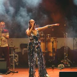 Im Vordergrund steht eine Sängerin im glitzernden Outfit. Sie hält das Mikrofon an ihren Mund und scheint den anderen Arm über ihrem Kopf zu schwingen. Im Hintergrund spielt ein Mann Saxophon.