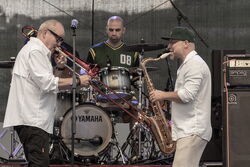 Drei Musiker, hinten der Schlagzeuger, links davor ein Posaunenspieler und rechts, ein Mann der Saxophon spielt.