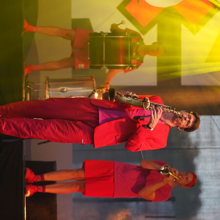 Das Bild zeigt drei Männer in roter Kleidung mit Instrumenten. Einer mit Trommel, einer mit Saxophon und der dritte mit Trompete.