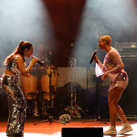 Das Bild zeigt zwei Sängerinnen, die sich anschauen und zueinander singen. Im Hintergrund sind weitere Musiker.