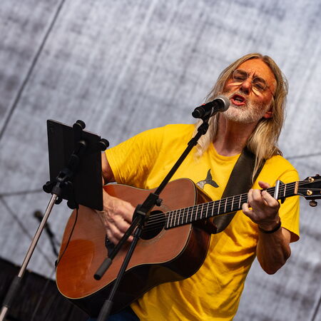 Das Foto zeigt einen Mann mit langem Haar, gelben Oberteil und einer Gitarre. Er spielt Gitarre und singt gleichzeitig in das Mikrofon vor ihm.