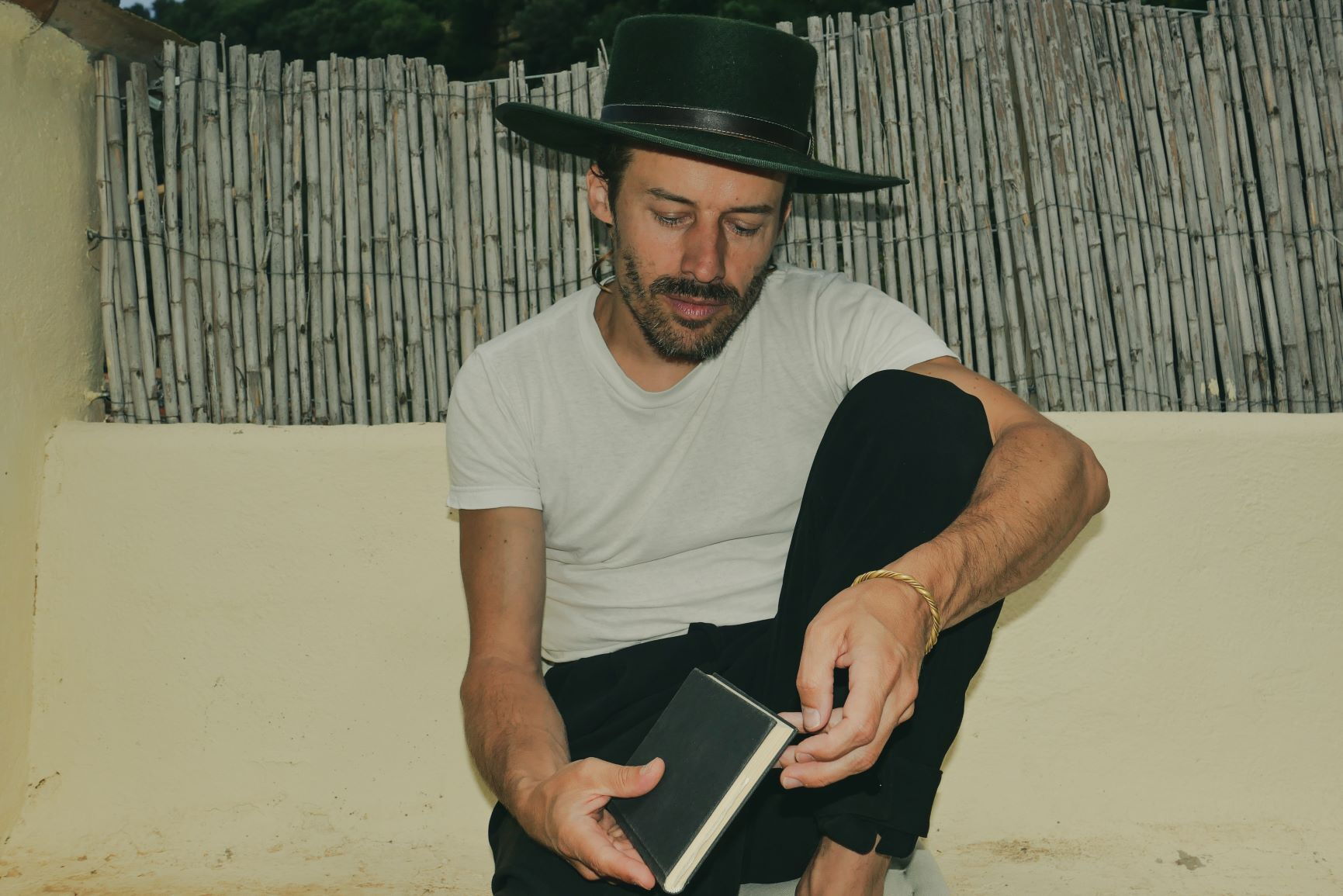 Bonaparte, ein Mann mit dunklem Hut und Vollbart, sitzt entspannt vor einem Bambuszaun auf einer beigen Terrasse. Er trägt ein weißes T-Shirt und schwarze Hosen, hält ein kleines schwarzes Notizbuch in den Händen und scheint darin zu blättern.