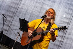 Das Foto zeigt einen Mann mit langem Haar, gelben Oberteil und einer Gitarre. Er spielt Gitarre und singt gleichzeitig in das Mikrofon vor ihm.