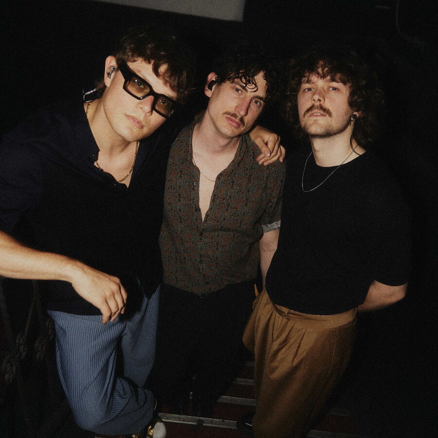 Drei junge Männer mit lockigem Haar stehen eng beieinander in einem dunklen Raum. Der Mann links trägt eine Sonnenbrille und ein dunkles Hemd, der mittlere ein gemustertes, leicht geöffnetes Hemd, der rechte ein schwarzes T-Shirt mit einer Halskette.