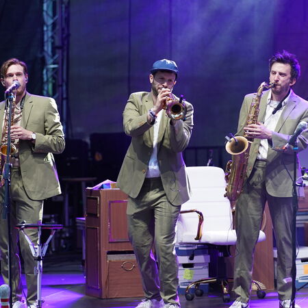 Drei Männer, welche alle den gleichen grünen Anzug tragen. Die beiden äußeren spielen Saxophon der in der Mitte Trompete.
