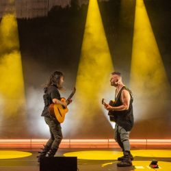 Die beiden Musiker stehen einander zugewand und spielen Gitarre. Das Licht der Scheinwerfer ist gelb.