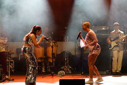 Das Bild zeigt zwei Sängerinnen, die sich anschauen und zueinander singen. Im Hintergrund sind weitere Musiker.