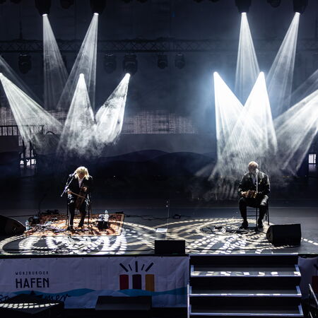 Zwei Musiker, welche mit etwas Abstand auf Stühlen sitzen. Es ist dunkel und sie werden jeweil von Scheinwerfern beleuchtet, die dann auch noch ein Muster auf den Boden werfen.