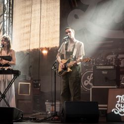 Zwei Musiker auf der Bühne, links eine Frau am Keyboar, rechts ein Mann an der Gitarre. Beide haben ein Mikrofon vor sich stehen und singen. Das Bild ist seitlich aufgenommen.