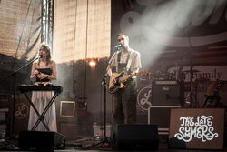 Zwei Musiker auf der Bühne, links eine Frau am Keyboar, rechts ein Mann an der Gitarre. Beide haben ein Mikrofon vor sich stehen und singen. Das Bild ist seitlich aufgenommen.