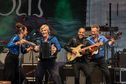 Es sind 4 Musiker auf der Bühne abgebildet, sie alle sind am lachen und musizieren. Das ganze Bild strahlt eine fröhliche, lebendige Atmosphäre aus.