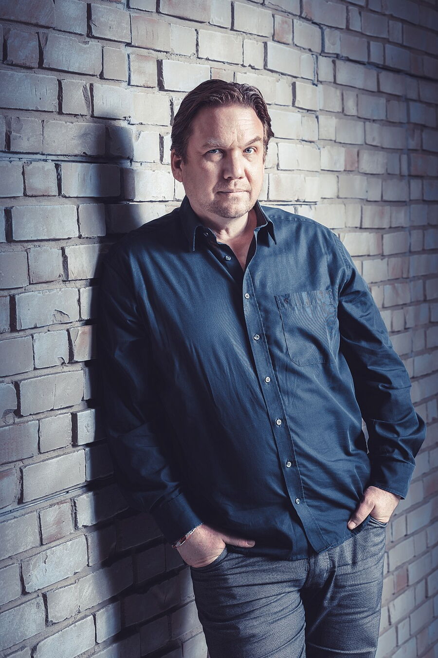 Matthias Brodowy lehnt lässig an einer grauen Backsteinmauer, die Hände in die Hosentaschen gesteckt und den Blick in die Kamera gerichtet. Er trägt ein blaues Hemd und wird von hellem Scheinwerferlicht bestrahlt.