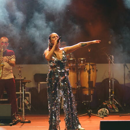 Im Vordergrund steht eine Sängerin im glitzernden Outfit. Sie hält das Mikrofon an ihren Mund und scheint den anderen Arm über ihrem Kopf zu schwingen. Im Hintergrund spielt ein Mann Saxophon.