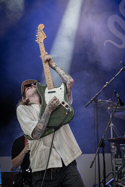 Der fotografierte Gitarrist hält seine Gitarre senkrecht in die Höhe während er darauf spielt. Auch sein Kopf liegt im Nacken, er schaut in die Luft.