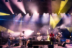 Das Bild zeigt 6 Musiker auf der Bühne, alle mit ihren verschiedenen Instrumenten. Die Scheinwerfer gehen in alle möglichen Richtungen und strahlen in lila und gelb.