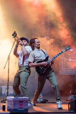 Auf dem Bild sind zwei Musiker in grünen Hosen welche sich nach hinten lehnen, sodass sich ihre Schultern berühren. Der Linke spielt Trompetem, der Rechte Gitarre.