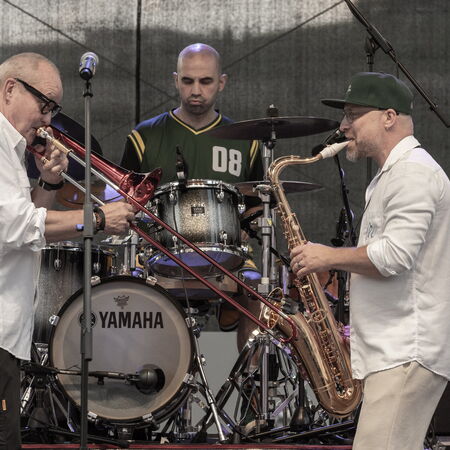 Drei Musiker, hinten der Schlagzeuger, links davor ein Posaunenspieler und rechts, ein Mann der Saxophon spielt.