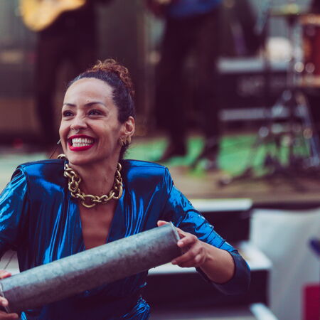 Ayom im blauen glänzenden Kleid, auffälliger, goldener Kette und einem breiten Grinsen. Sie befindet sich vor einer Bühne und trägt etwas in ihren Händen.