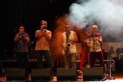 Das Bild zeigt die Band in einem sehr dunklen Licht mit Nebel. Alle 4 blicken nach vorne und spielen ein Blasinstrument. Im Hintergrund ist ein weiterer Mann versteckt.