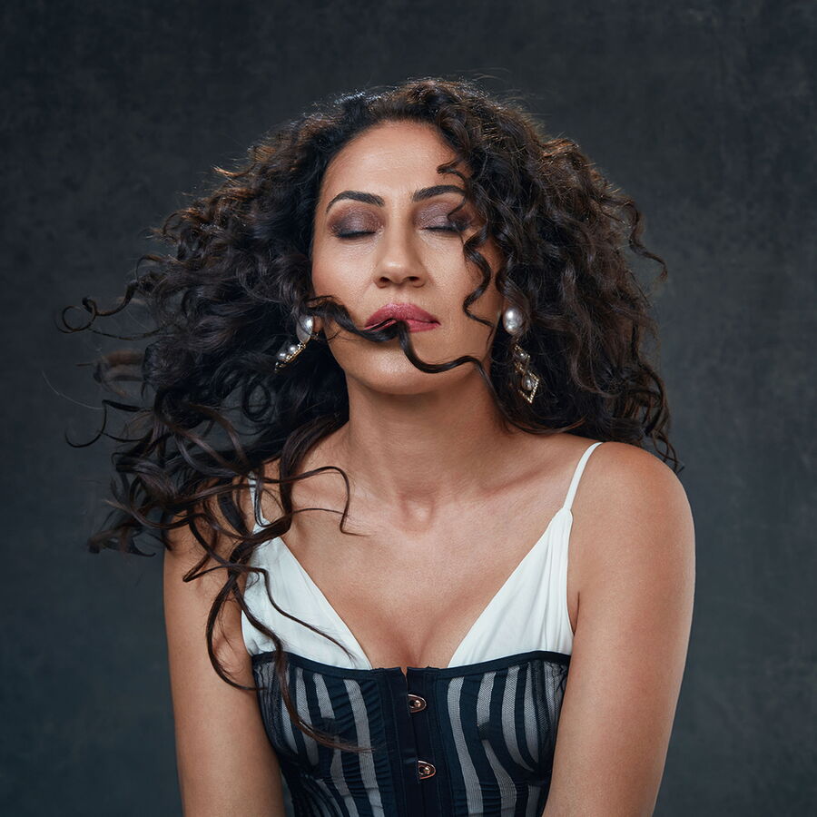 Das Bild zeigt Aynur mit geschlossenen Augen und wehendem, lockigem Haar. Sie trägt ein ärmelloses Oberteil mit schwarzen und weißen Streifen und auffällige Ohrringe mit Perlen- und metallischen Details. Der Hintergrund ist dunkel und neutral, was den Fok