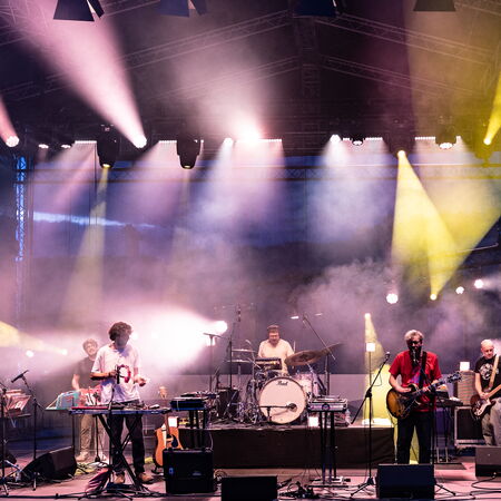 Das Bild zeigt 6 Musiker auf der Bühne, alle mit ihren verschiedenen Instrumenten. Die Scheinwerfer gehen in alle möglichen Richtungen und strahlen in lila und gelb.