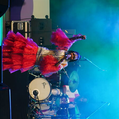Sängerin in aufälligem rot-goldenen Kleid beim singen mit dem linken arm in der Höhe als würde sie tanzen.