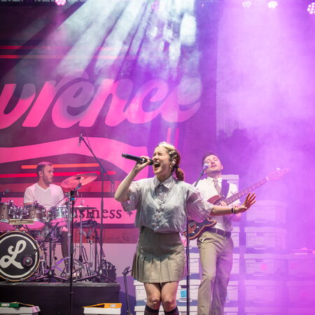 Das Bild zeigt drei Bandmitglieder in Action. Im Vorderund eine Sängerin mit Mikro in der Hand. Im Hintergrund Gitarrist und Schlagzeuger.