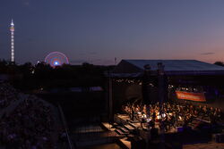 Das Bild zeigt eine Aufnahme der Bühne von oben. Es ist bereits dunkel. Das Orchester ist jecoh hell beleuchtet. Im Hintergrund sieht man beleuchtete Fahrgeschäfte des Volksfestes.