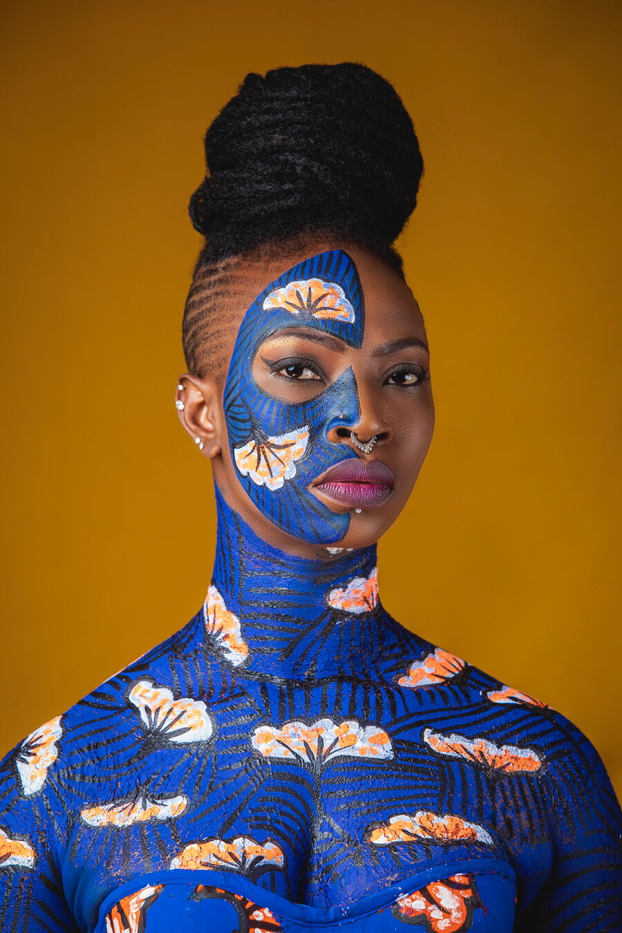 Das Bild zeigt Dobet Gnahore von der Schulter aufwärts vor einem braunen Hintergrund. Sie ist am ganzen Körper, bis auf die rechte Gesichtshälfte, blau angemalt mit Mustern des Bureka Africana, ein Baum in Afrika. Sie schaut direkt in die Kamera.