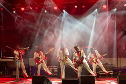 Das Bild zeigt eine Band von 6 personen alle spielen ein Instrument und sie stehen auf einer Bühne. 4 Männer sind in der gleichen Pose, un welcher sie auf dem rechten Bein stehen und das Linke in die Luft heben.