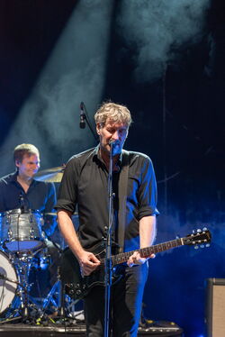 Sänger der Band an Gitarre und Mikrofon, im Hintergrund der Schlagzeuger.