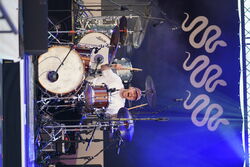 Schlagzeuger in Action. Er hält beide Drumsticks nach oben und sein Mund ist weit aufgerissen, so als würde er mitsingen.