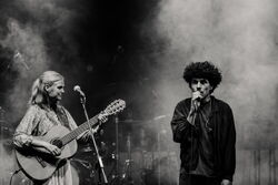 Das schwarz-weiß Bild zeigt links eine Frau mit Gitarre. Rechts einen Mann mit Mikrofon, welcher gerade singt. Die Atmospähre ist durch Nebel sehr mystisch.
