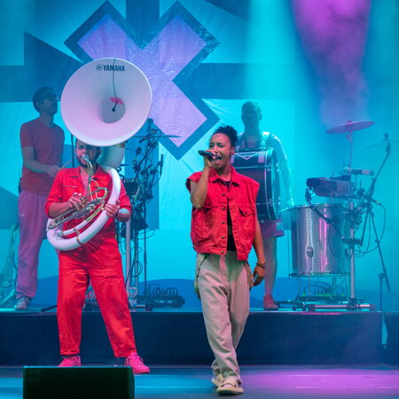 Die Musiker der Band im Einsatz bei lila Licht. Links ein Mann mit Sousaphon und rechts eine singendePerson die den linken Arm nach oben streckt.