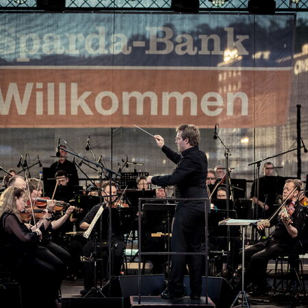 Das Bild zeigt ein Orchester mit ihrem Dirigenten vorne dran. Sie stehen auf der Bühne des Hafen Sommers.