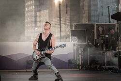 Ein Mann in grauer Hose und schwarzem Oberteil, welcher leidenschaftlich Gitarre spielt.