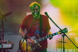 Das Bild zeigt einen Mann mit Brille, welcher Gitarre spielt und dazu singt. Er trägt ein rotes Oberteil mit blauer Aufschrift.
