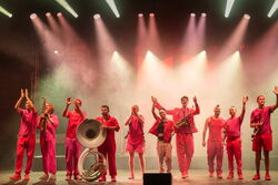 Das Bild zeigt die gesamte Band, 10 Personen. Alle tragen rote Kleidung und sind in einer Reihe aufgestellt. Sie winken und klatschen.