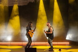 Die beiden Musiker stehen einander zugewand und spielen Gitarre. Das Licht der Scheinwerfer ist gelb.