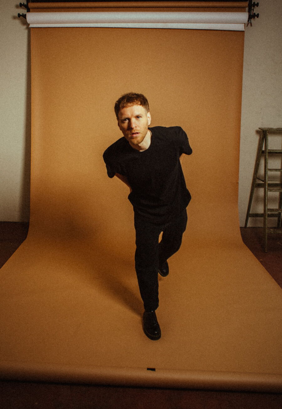 Ein junger Mann mit roten Haaren und Bart trägt ein schwarzes Outfit und steht vor einem warmbraunen Fotohintergrund. Er beugt sich leicht nach vorne, mit den Armen hinter dem Rücken verschränkt. Im Hintergrund rechts ist eine alte Holzleiter zu sehen.