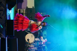 Sängerin in aufälligem rot-goldenen Kleid beim singen mit dem linken arm in der Höhe als würde sie tanzen.